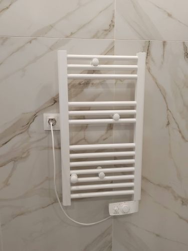 На стену ванной комнаты закреплен белый прибор с подключением к розетке. Устройство установлено на плитку, подключено к электросети, готово к использованию.