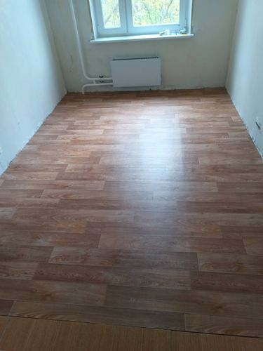 На полу ровно уложен линолеум с текстурой дерева, без видимых швов и дефектов. Поверхность гладкая, плотно прилегает к основанию, создавая аккуратный и завершенный вид.