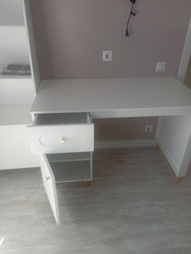 Собран белый стеллаж с полками, ящиком и дверцей. Установлены ножки, закреплены детали. Готовая конструкция стоит на светлом полу, рядом розетка на стене.