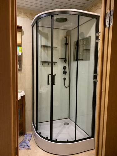 Установлена современная душевая конструкция с прозрачными стенками и черными рамами. Поддон белый, смеситель и лейка закреплены, полки для принадлежностей размещены внутри.