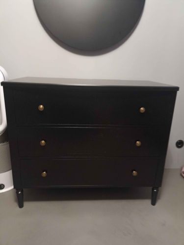 Собран новый черный комод с тремя ящиками и золотыми ручками, установлен на ровной поверхности, все детали надежно закреплены, конструкция устойчива, готов к использованию.