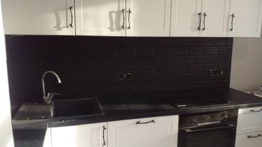 На изображении показана кухня с черной плиткой на стене. Плитка уложена ровно, швы аккуратно заполнены, создавая стильный контраст с белыми шкафами и черной столешницей.