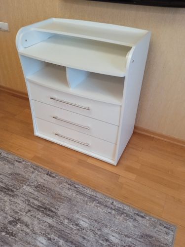 Собран белый комод с тремя ящиками и двумя полками. Установлены ручки, закреплены боковые панели и задняя стенка. Проверена устойчивость и функциональность всех элементов.