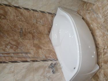 Ванная комната с новой белой ванной, установленной на кафельном полу. Стены облицованы плиткой с мраморным узором. Смеситель и душевой шланг закреплены на стене.