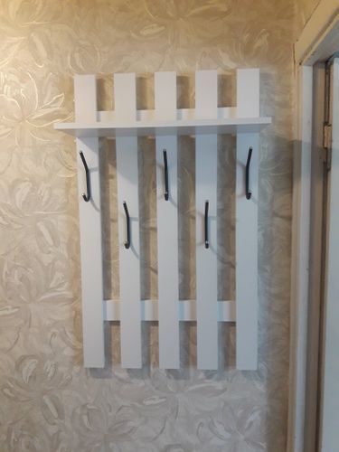 На стене закрепили белую полку с пятью крючками. Конструкция надежно установлена на декоративной поверхности, обеспечивая функциональность и эстетичность в интерьере.