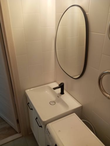 Изображение ванной комнаты с белой раковиной, установленной на тумбе, черным смесителем и овальным зеркалом на стене. Рядом расположена стиральная машина.