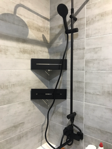 Изображение ванной комнаты с установленным душем. На стене закреплены черные полки и душевая система. Видны плитка и элементы сантехники.