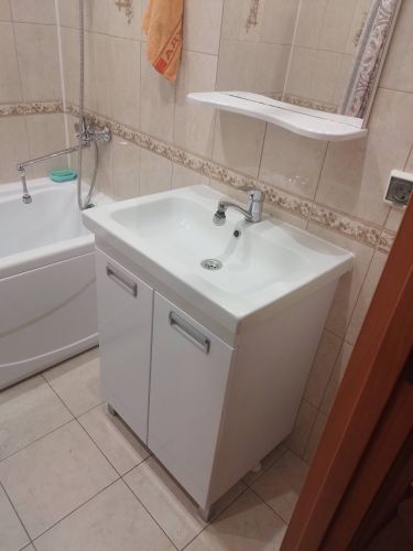 Изображение ванной комнаты с установленной новой раковиной и шкафчиком. Видна ванна с душем, плитка на стенах и полу, полка над умывальником, полотенце.