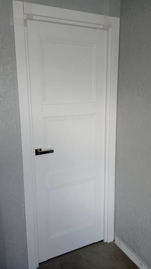 Установлена новая белая дверь с ручкой, аккуратно закреплена в проеме, добавлены наличники, обеспечивающие плотное прилегание. Завершен монтаж, обеспечена функциональность и эстетика.