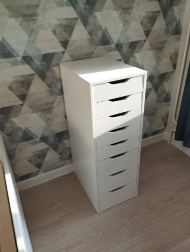 Собран белый комод с шестью выдвижными ящиками, установлен в углу комнаты на светлом полу, на фоне геометрических обоев. Все элементы надежно закреплены, конструкция устойчива.