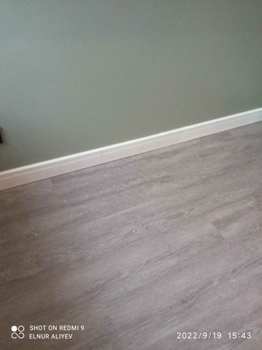 Установлен новый белый плинтус вдоль стены, аккуратно прилегающий к серому полу, создавая завершенный и эстетичный вид интерьера.