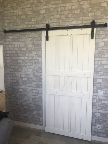 Установлена белая раздвижная створка на черном металлическом направляющем механизме. Дверь плавно перемещается вдоль стены с серыми кирпичными обоями, обеспечивая удобство и стиль.