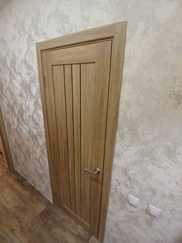 Установлена деревянная створка с петлями, ручкой и наличниками. Полотно ровно закреплено в проеме, обеспечивая плавное открытие и закрытие. Завершены отделочные работы вокруг конструкции.