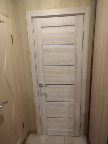 Изображена установленная светлая дверь с ручкой и петлями в проеме. Завершены монтажные работы, включая крепление полотна и регулировку конструкции.