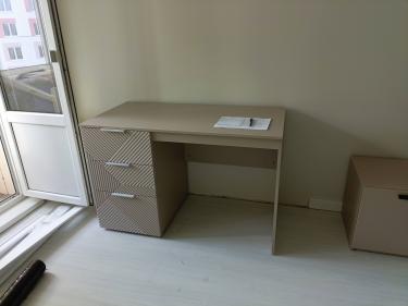 Собрана новая мебель с тремя ящиками и гладкой столешницей. Все детали аккуратно соединены, обеспечивая устойчивость и функциональность. Установлена в помещении у окна.