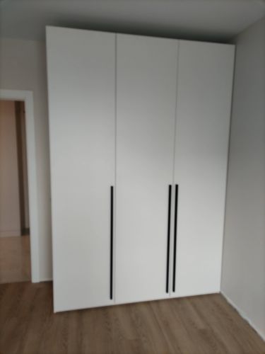 Собран вместительный шкаф с тремя дверцами, аккуратно установлен в углу комнаты. Белая поверхность гармонирует с интерьером, черные ручки добавляют стильный акцент.