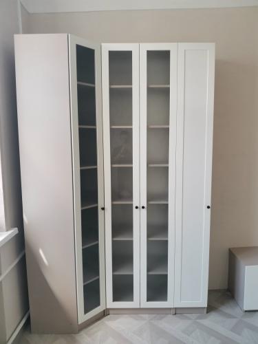 Собран шкаф с белыми дверцами и стеклянными вставками, установлены полки, закреплены ручки. Конструкция аккуратно размещена в углу комнаты, обеспечивая функциональность и стиль.