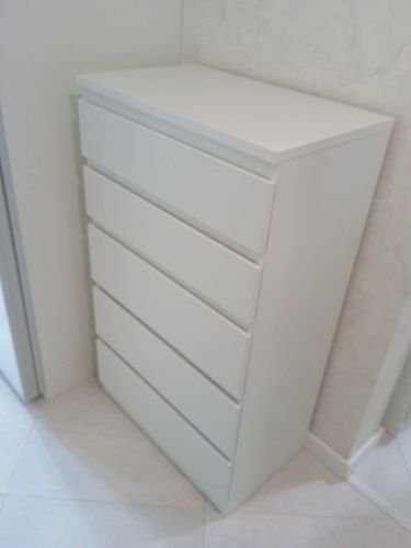 Собран белый комод с пятью ящиками, установлен в углу комнаты. Все детали соединены, фасады ровные, конструкция устойчива, ящики выдвигаются плавно.