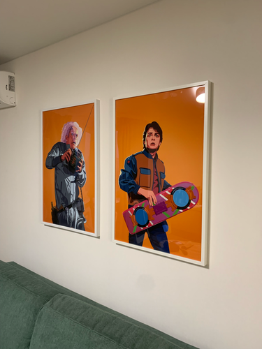 Изображены две яркие картины в белых рамках, аккуратно закрепленные на светлой стене над зеленым диваном.