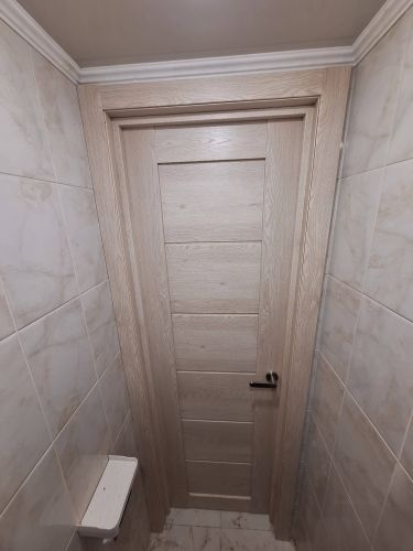 Установлена светлая дверь с ручкой, аккуратно встроена в проем, обрамлена наличниками, плотно прилегает к стенам, гармонично сочетается с плиткой, завершает интерьер комнаты.