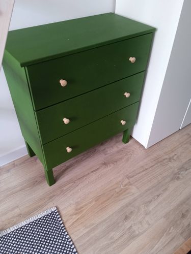 Собран зелёный комод с тремя ящиками и деревянными ручками, установлен на ровный пол. Все детали соединены, конструкция устойчива, ящики выдвигаются плавно.