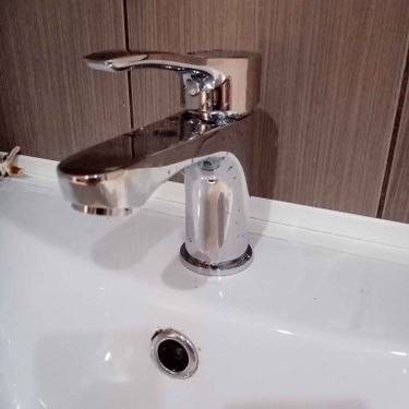 Изображен блестящий смеситель, установленный на белоснежной раковине, с аккуратно закрепленными элементами. Фон выполнен в тёплых коричневых тонах.