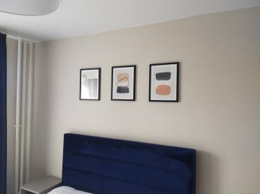 Изображение пустой стены с тремя картинами в рамках над темно-синей спинкой кровати. Ковер не виден.