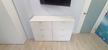 Собран белый комод с шестью ящиками, установлен в комнате. Все детали соединены, ручки прикреплены, конструкция устойчива, поверхность ровная. Работа завершена аккуратно и качественно.