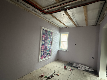 Стены оклеены светлыми обоями, создающими ровную поверхность. В комнате установлено окно, добавляющее естественное освещение. На полу лежат инструменты и остатки материалов.