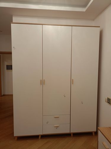 Собран шкаф с тремя дверцами и двумя ящиками. Установлены ручки, выровнены панели, закреплены ножки. Конструкция устойчива, готова к использованию.