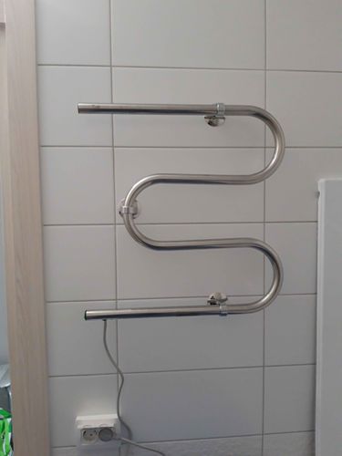 На стене ванной комнаты закреплен металлический прибор, подключенный к электросети. Устройство надежно зафиксировано, кабель аккуратно подведен к розетке.