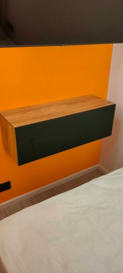 Изображен интерьер с яркой стеной, на которой закреплен деревянный шкаф. На переднем плане виден край кровати с белым покрывалом.