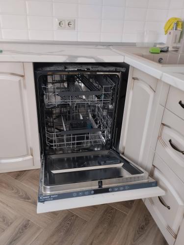 Изображение показывает кухню с открытой посудомоечной машиной, встроенной в мебель. Видны деревянный пол и белая столешница. На заднем плане розетки и кухонные принадлежности.