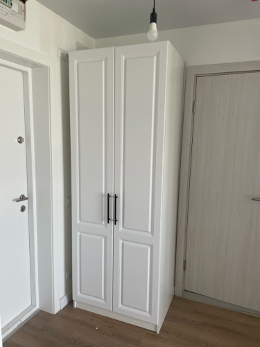 Собран белый шкаф с двумя дверцами, установлен в углу комнаты. Закреплены ручки, отрегулированы петли. Проверена устойчивость и функциональность конструкции.