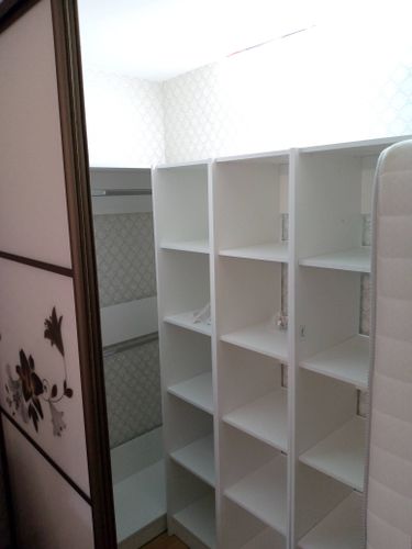 Белый стеллаж установлен в углу комнаты. Полки ровно закреплены, конструкция устойчивая. Пространство организовано для хранения вещей. Завершенная работа гармонично вписывается в интерьер.