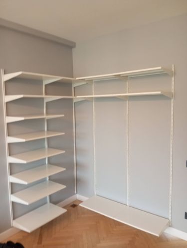 Установлены белые полки на стену в углу комнаты. Конструкция закреплена надёжно, обеспечивая устойчивость и функциональность. Полки ровные, идеально подходят для хранения вещей.