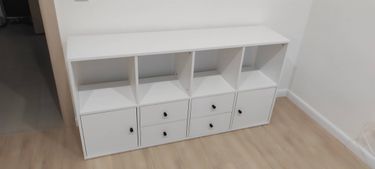 Собран белый стеллаж с полками и ящиками. Конструкция установлена на полу, все элементы аккуратно закреплены, дверцы и ручки на месте. Работа выполнена качественно и надежно.