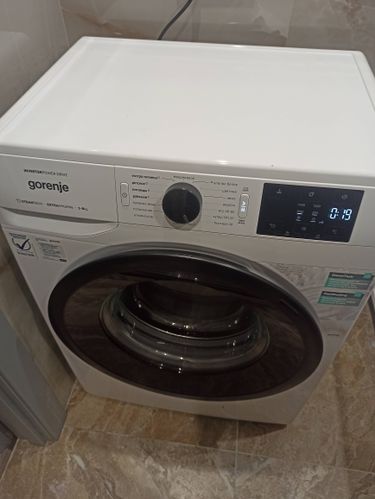 Изображена стиральная машина Gorenje, установленная на плиточном полу. Видна панель управления с различными режимами стирки и дисплеем. Машина подключена к электросети.
