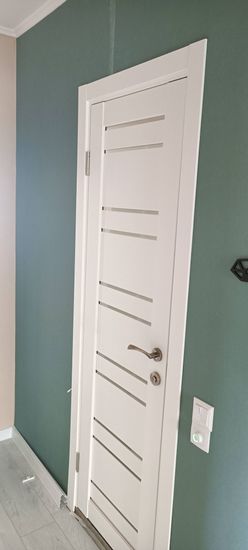 Установлена новая белая дверь с горизонтальными вставками, закреплена на петлях, добавлена ручка с замком. Оформлены наличники, конструкция выровнена, обеспечена плавная работа.