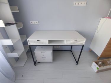 Собран белый стеллаж с несколькими полками, установлен в углу комнаты. Все детали аккуратно соединены, конструкция устойчива и готова к использованию.