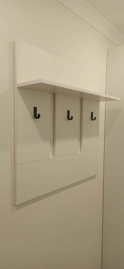На стене закреплена белая панель с тремя черными крючками и полкой сверху. Конструкция надежно установлена, обеспечивая удобное хранение одежды и аксессуаров.