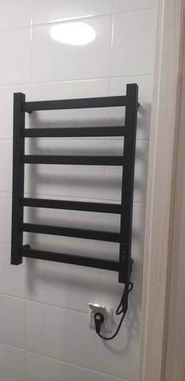 На стене ванной закреплён электрический прибор для сушки полотенец. Подключение выполнено к розетке, кабель аккуратно уложен. Устройство готово к использованию.