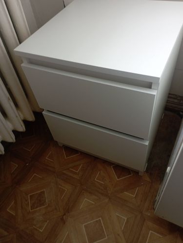 Собрана белая тумба с двумя выдвижными ящиками, установлена на ровный пол. Все детали аккуратно соединены, конструкция устойчива и готова к использованию.