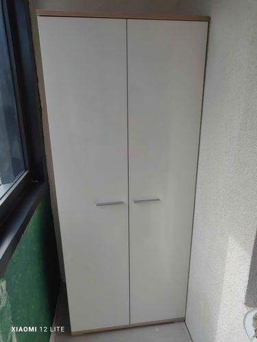 Изображение показывает установленный шкаф на балконе. Белые двери с ручками, аккуратно размещены у стены. Рядом окно, свет падает на поверхность.