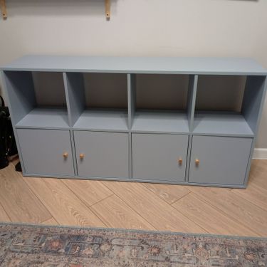 Собран аккуратный стеллаж с четырьмя открытыми секциями и тремя закрытыми отделениями. Конструкция установлена на полу, двери оснащены деревянными ручками.