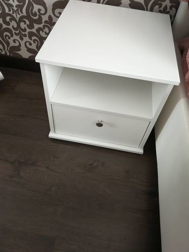 Собран белый шкафчик с полкой и ящиком, установлен на полу. Все детали соединены, ручка прикреплена, конструкция устойчива. Мебель готова к использованию, размещена в интерьере.