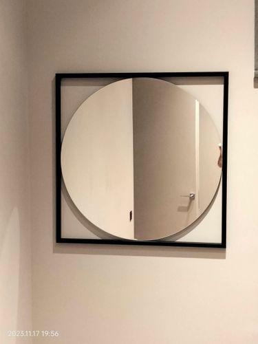 На стене закреплено круглое зеркало в квадратной раме. Оно висит ровно, надежно, гармонично вписываясь в интерьер. Работа выполнена аккуратно и профессионально.