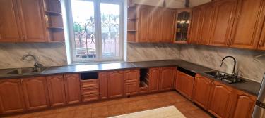 Изображена кухня с деревянными шкафами, двумя мойками, варочной панелью, мраморным фартуком и окном с решеткой. Светлая плитка на полу, современный стиль.