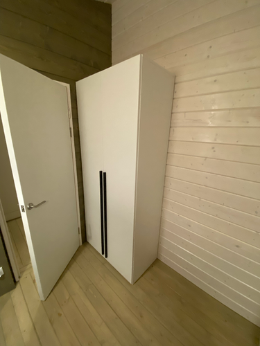 Изготовлен и установлен шкаф в углу комнаты. Белая поверхность, черные ручки. Двери закрыты. Пол деревянный, стены светлые. Рядом открытая дверь.