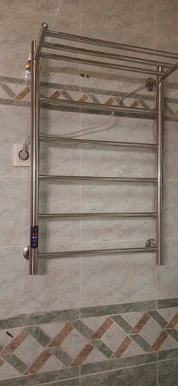 На стене ванной комнаты закреплен металлический прибор, подключенный к розетке. Устройство надежно установлено, кабель аккуратно спрятан, обеспечена безопасная эксплуатация.